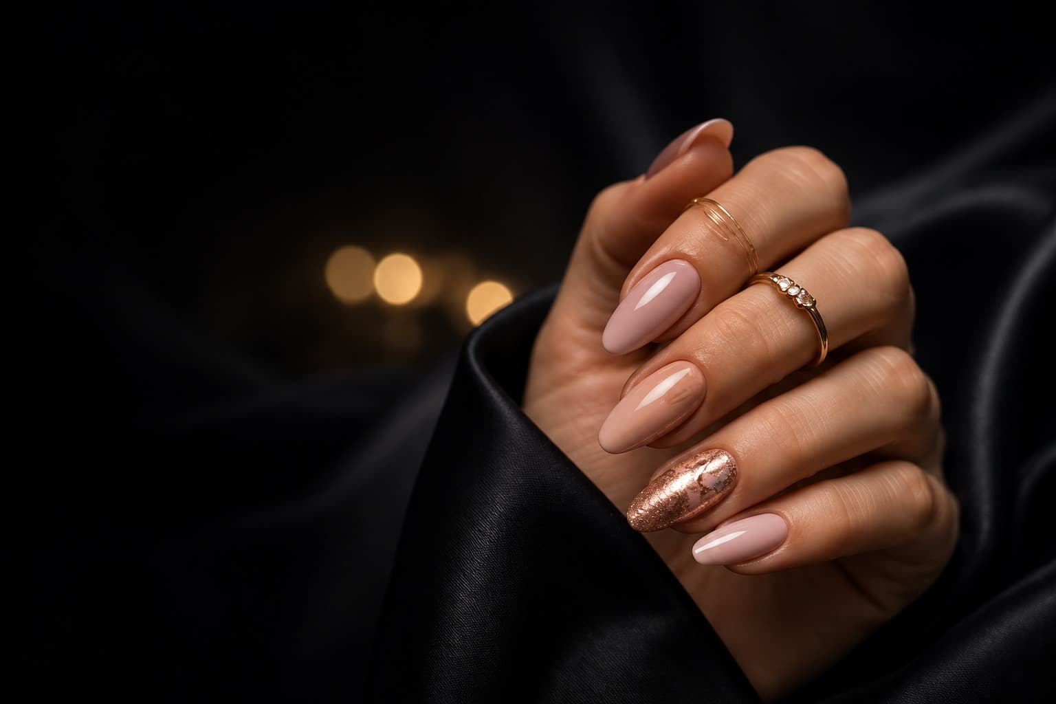 Hand with almond nails in nude pink and one rose-gold foil accent nail, wearing two gold rings